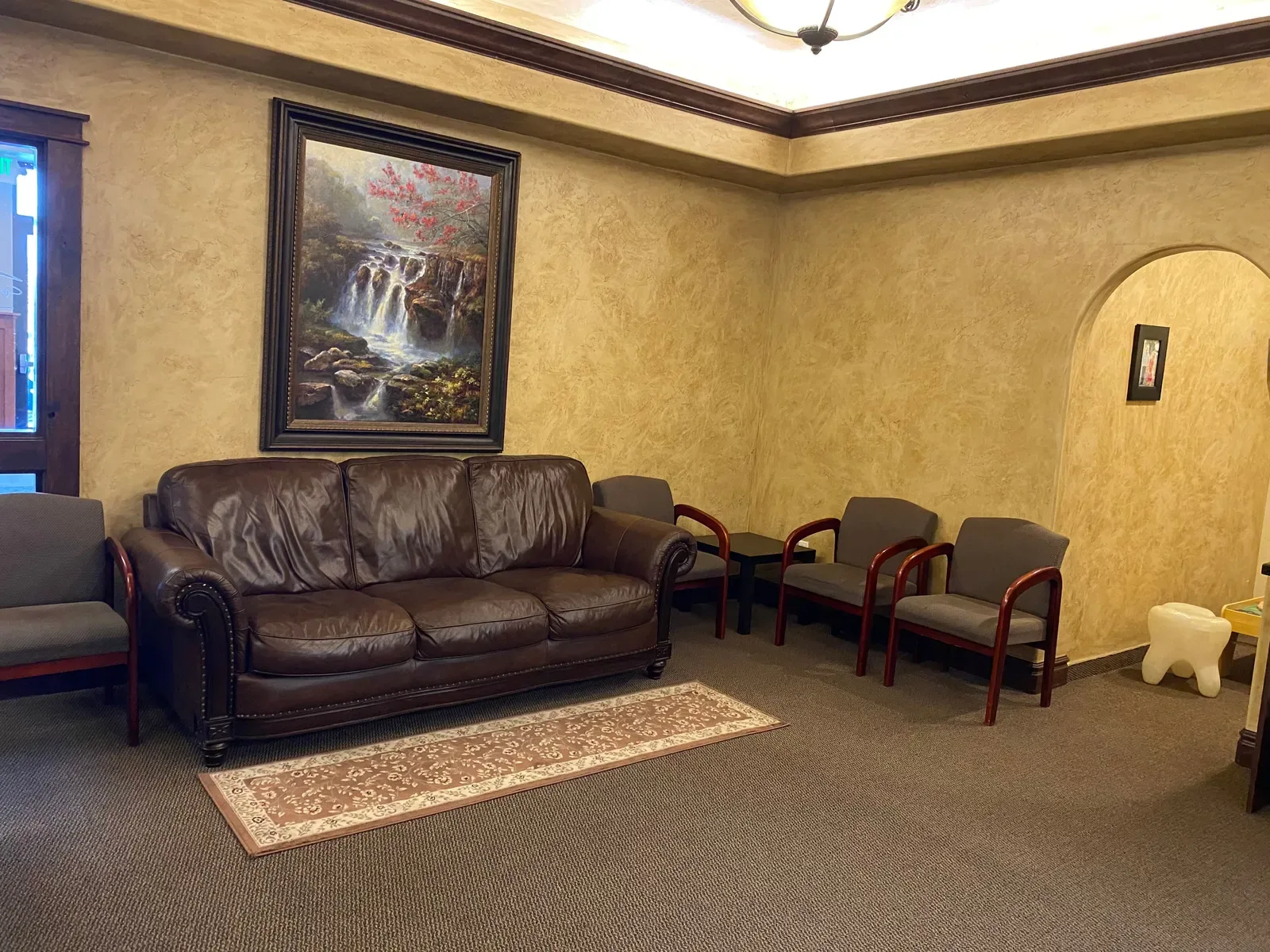 Waiting room with a brown leather sofa, armchairs, patterned rug, and a framed waterfall painting.